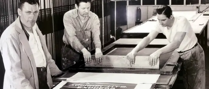 three men working on a large print in a factory, demonstrating the history of screen printing